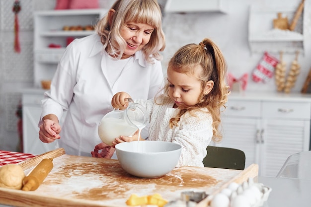 Füllt sich mit Milch Ältere Großmutter mit ihrer kleinen Enkelin kocht Süßigkeiten für Weihnachten in der Küche