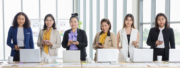 Fünf asiatische Geschäftsfrauen in formellen Anzügen, die im Büro zusammenstehen und Smartphones hinter einem hölzernen Konferenztisch mit Laptop-Diagrammen und Diagrammen darauf verwenden.