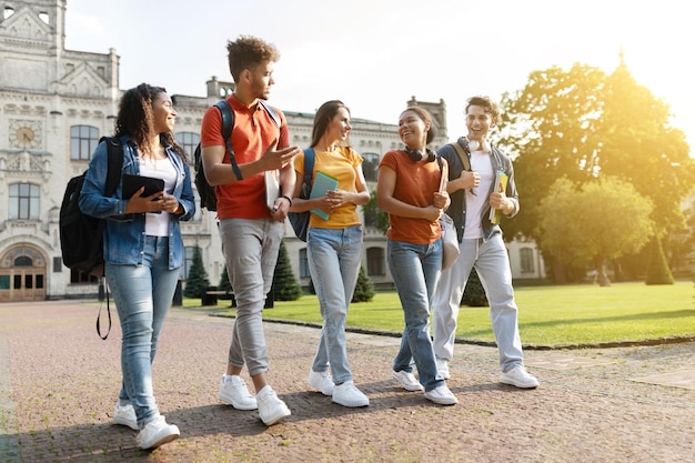 Fünf multiethnische Studenten unterhalten sich und lachen, während sie gemeinsam auf dem Universitätscampus spazieren gehen