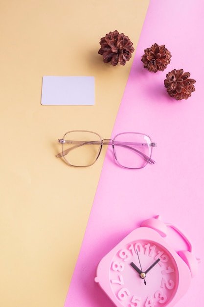 Fünfeckige Brille auf dem Foto im minimalistischen Sommerstil auf pastellrosa und gelbem Hintergrund. Wecker , Kiefernblüten , Visitenkarten