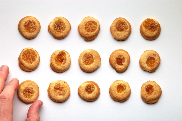 Fünfzehn handgemachte Kekse mit Aprikosenmarmelade in gleichmäßigen Reihen. Mann Hand nimmt eine von unten. isoliert auf weißem Hintergrund