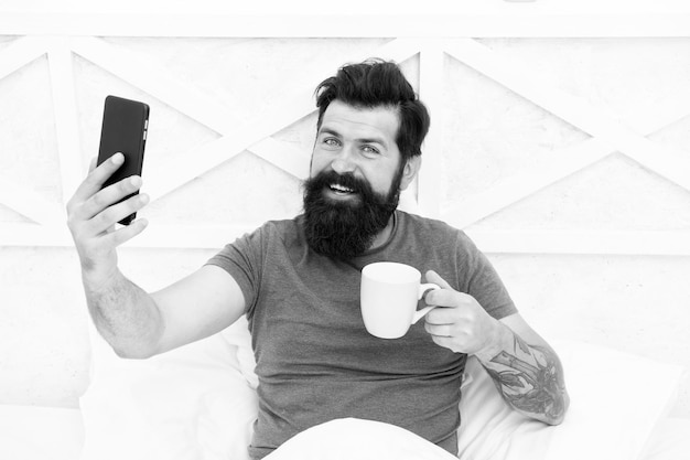 Fuente de energía Hombre barbudo disfruta de videoconferencias y café en la cama Hipster feliz sostiene una taza y un teléfono móvil Bebida energética caliente Aumento de los niveles de energía Comunicación energética Aumento de energía