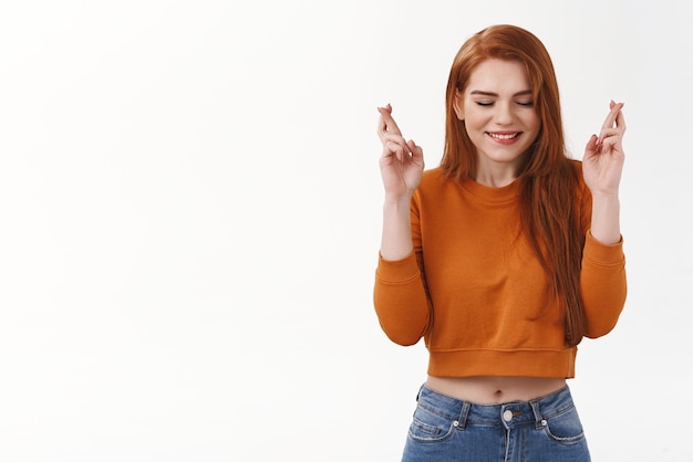 Für den Erfolg beten Zuversichtlich, verlockend, niedliche rothaarige Frau, die Make-up trägt, abgeschnittener, stilvoller Pullover, Augen schließen, lächelnd, optimistisch, Aufregung, gekreuzte Finger, viel Glück, glauben, dass ein Traum wahr wird, wünschen