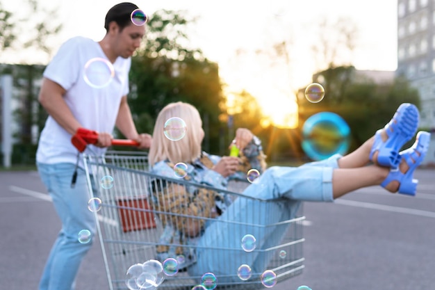 Foto fuera de la imagen enfocada de la feliz pareja joven diviértete con un carrito de la compra y sopla burbujas en el estacionamiento de un supermercado.