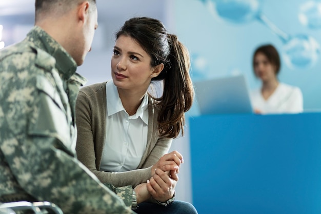 Fürsorgliche Frau, die Händchen hält und ihren Militärmann unterstützt, während sie mit ihm auf dem Krankenhausflur spricht