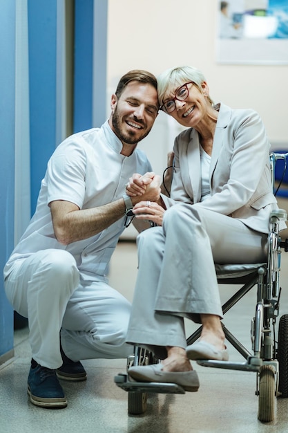 Fürsorglicher Arzt und seine ältere Patientin im Rollstuhl halten sich an den Händen in einer Lobby der Klinik