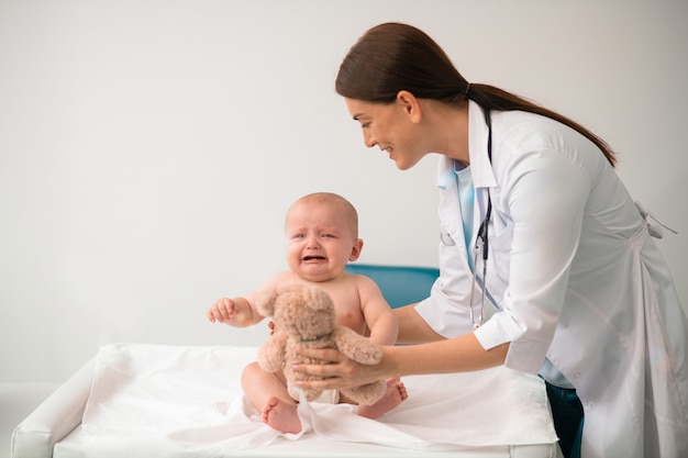 Fürsorglicher Kinderarzt, der einem verärgerten Baby einen Teddy schenkt