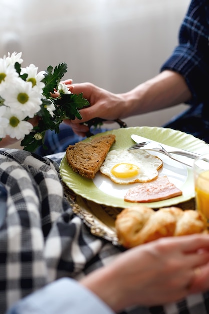 Fürsorglicher Mann brachte Frühstück mit Eiern, Speck, Toast und Orangensaft, Blumen in das Bett seiner Freundin, das mit Plaid bedeckt ist