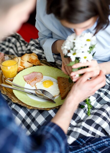 Fürsorglicher Mann brachte Frühstück mit Eiern, Speck, Toast und Orangensaft, Blumen in das Bett seiner Freundin, das mit Plaid bedeckt ist