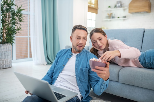 Fürsorglicher Vater, der mit einem Laptop auf dem Boden sitzt und seiner Tochter einen Teller mit Marshmallows hinhält