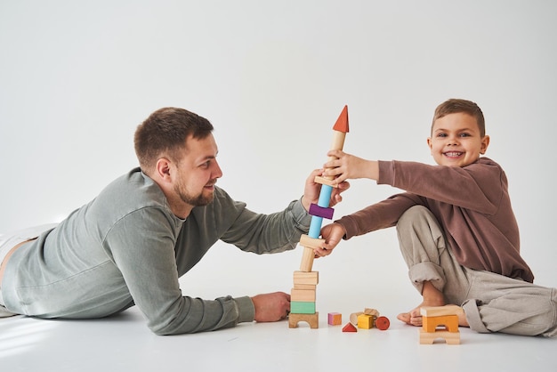 Foto fürsorglicher vater hilft seinem sohn, auf dem boden auf weißem hintergrund zu spielen vater und kind bauen turm aus bunten holzziegeln und haben gemeinsam spaß