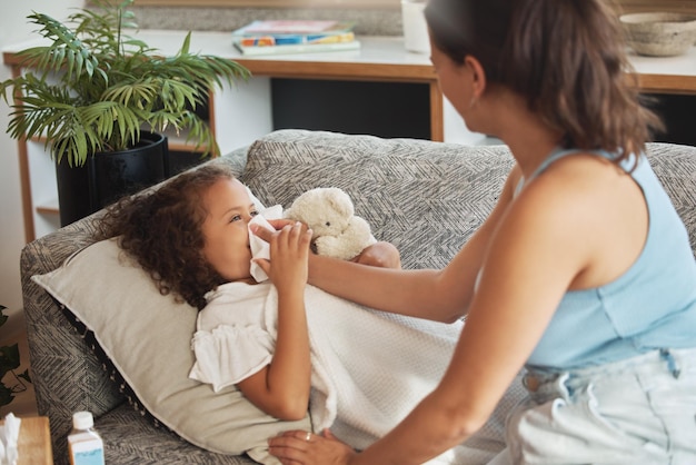 Fürsorgliches, liebevolles und besorgtes Elternteil, das sich um krankes Kind kümmert, das an kalter Grippe oder Covid-Virus leidet Tochterkind und krankes Mädchen liegen auf dem Sofa, während Mutter ihr hilft, ihre Nase im heimischen Wohnzimmer zu putzen