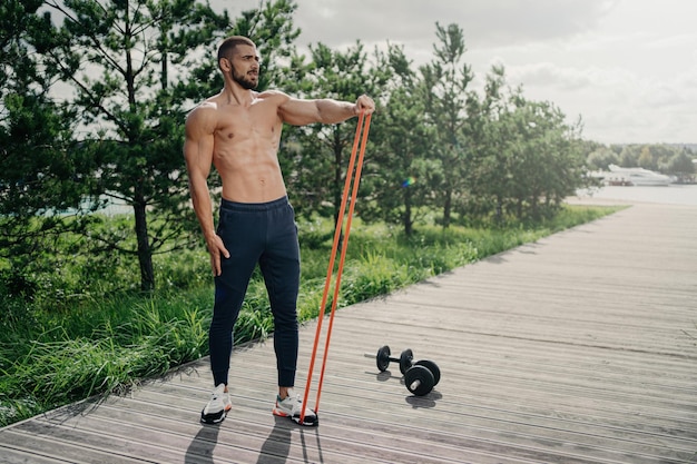 Fuerte atleta barbudo usa banda de resistencia para entrenamiento al aire libre realiza ejercicios con equipo deportivo