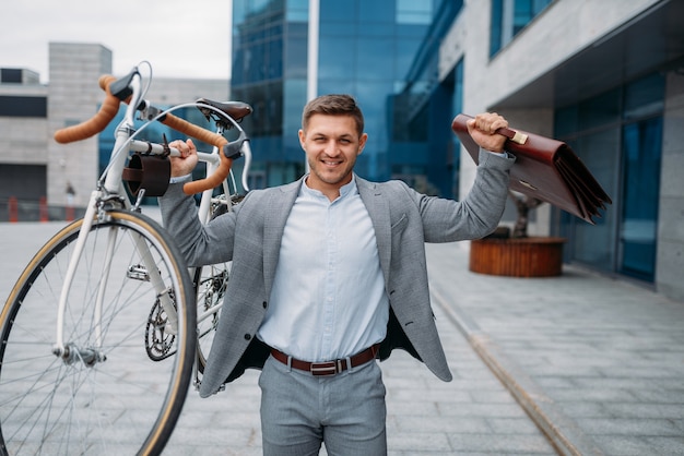 Fuerte hombre de negocios tiene bicicleta y maletín en el edificio de oficinas de cristal en el centro de la ciudad. Persona de negocios en transporte ecológico en las calles de la ciudad, estilo urbano