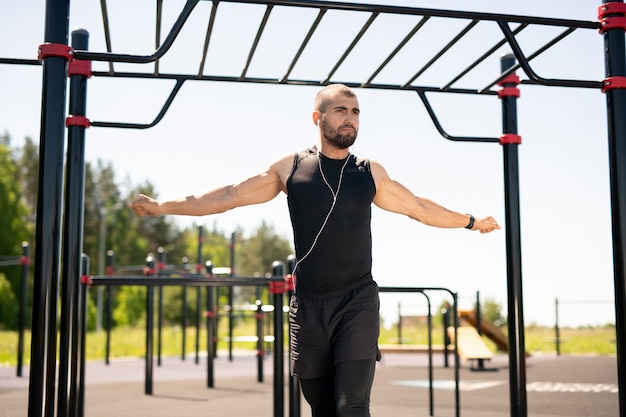 Fuerte joven atleta masculino en ropa deportiva negra extendiendo sus brazos mientras hace ejercicio en el campo de deportes al aire libre