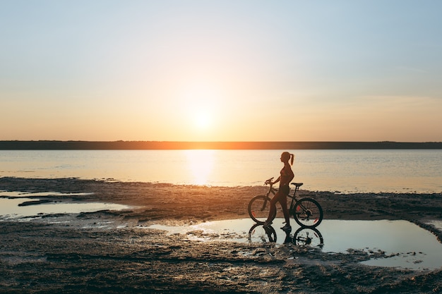 La fuerte mujer rubia con un traje colorido se encuentra cerca de la bicicleta en el agua al atardecer en un cálido día de verano. Concepto de fitness.