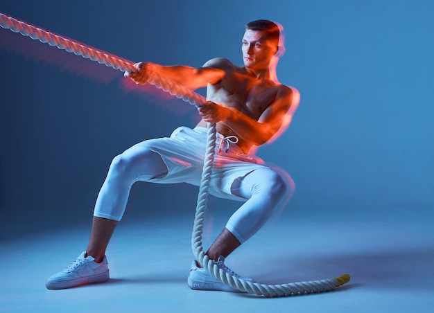 Fuerte musculoso apuesto joven sosteniendo la cuerda en ropa deportiva blanca sobre fondo azul con largo