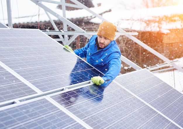 Fuerte técnico masculino en traje azul instalando módulos solares fotovoltaicos azules como fuente de energía renovable.