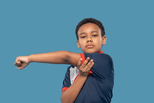 Fuerza, músculos. Grave niño afroamericano en edad escolar primaria el ejercicio de las manos haciendo ejercicios físicos sobre fondo azul.