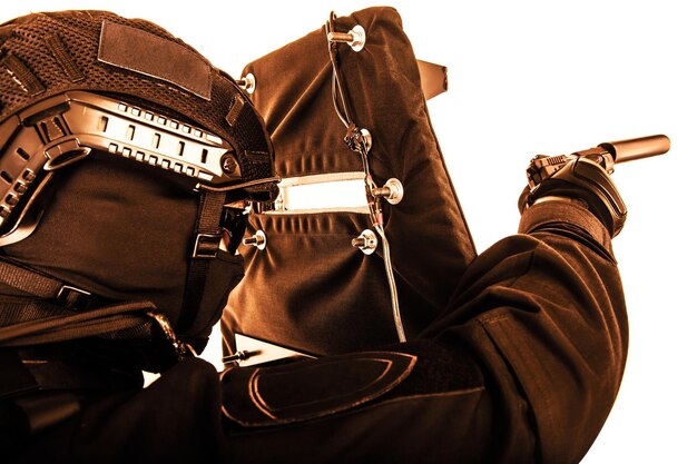 Fuerzas especiales de policía miembro del equipo de asalto de combate oficial del grupo táctico en uniformes negros apuntando con una pistola silenciada mientras se esconde detrás cubriéndose con un escudo balístico tonado coloreado