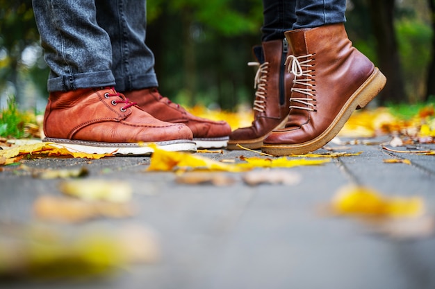 Füße eines verliebten Paares in den braunen Schuhen auf dem Weg des Herbstparks, gestreut mit gefallenen Blättern. Mädchen steht auf den Zehen. Kuss-Konzept