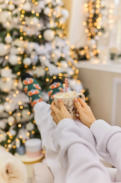 Füße in Weihnachtssocken neben dem Weihnachtsbaum. Frau sitzt an der Decke, trinkt heißes Getränk und entspannt sich beim Aufwärmen der Füße in Wollsocken. Winter- und Weihnachtsferienkonzept