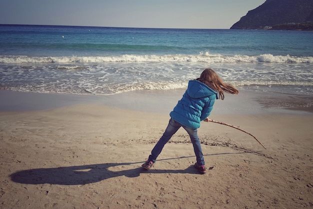 Full-Length-Rücksicht auf ein spielerisches Mädchen mit einem Stock am Strand gegen den Himmel
