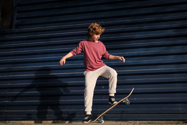 Full-Length-Skater in lässiger Kleidung macht einen Trick auf einem Skateboard gegen eine dunkelblaue Wand auf einer Stadtstraße