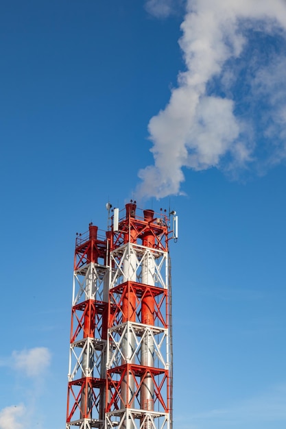 Fumaça nociva branca saindo dos canos vermelho e branco com antenas de comunicação móvel em uma fábrica no centro da cidade no contexto de um céu azul claro Problemas ambientais