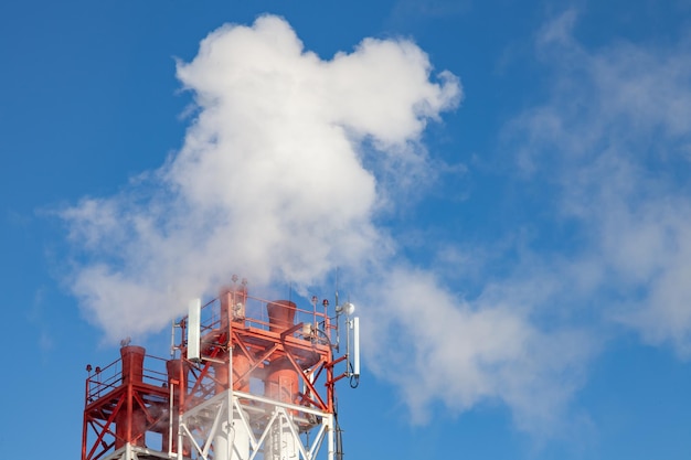 Fumaça prejudicial branca saindo dos canos vermelho e branco com antenas de comunicação móvel em uma fábrica no centro da cidade no contexto de um céu azul claro Problemas ambientais