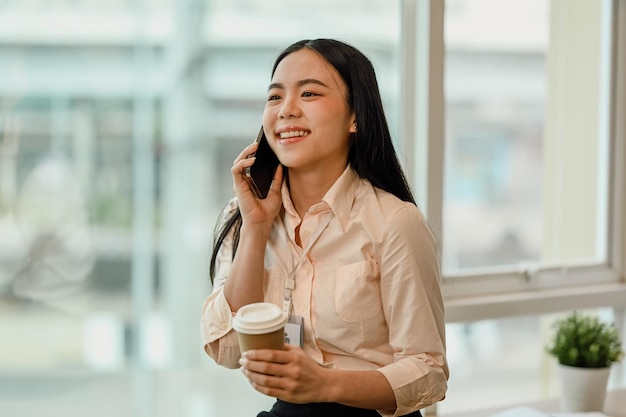 Funcionária asiática alegre falando no telefone celular perto da janela do escritório