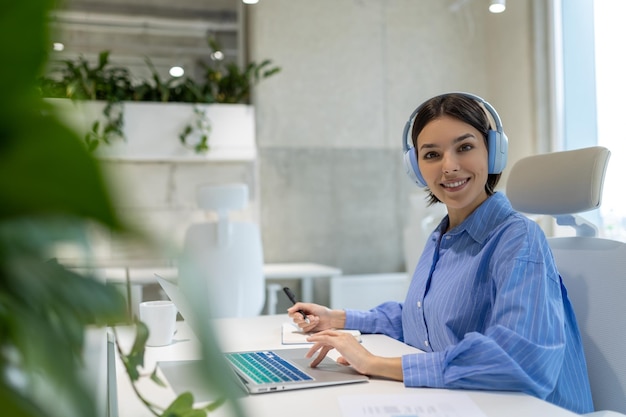 Funcionária de escritório alegre com fones de ouvido sentada no computador portátil fazendo anotações em seu caderno