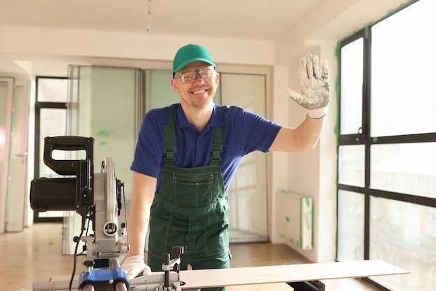 Funcionário acena com a mão em pé ao lado da máquina de serrar em sala reformada trabalhador feliz com equipamento