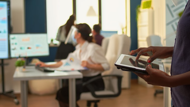 Funcionário africano usando tablet em pé na sala de escritório e equipe de analistas financeiros trabalhando em segundo plano. colegas de trabalho multiétnicas respeitando a distância social em empresas de negócios durante a pandemia global