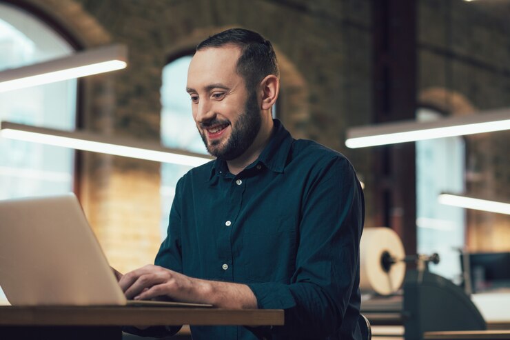 Funcionário alegre e entusiasmado sentado à mesa na editora e sorrindo enquanto usa um laptop moderno