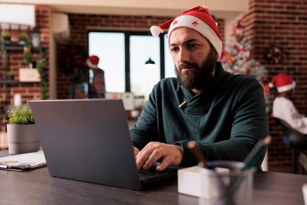 Funcionário da empresa com chapéu de papai noel trabalhando no laptop no escritório festivo, luzes da árvore de natal e decorações. Usando o computador no local de trabalho com enfeites de férias sazonais e decoração de Natal.