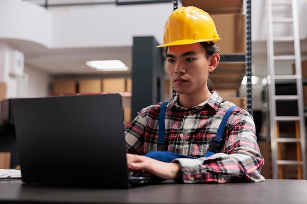 Foto funcionário de armazém asiático controlando estoque usando software de armazenamento de mercadorias no laptop. manipulador de pacotes jovens do depósito verificando o pedido on-line do cliente no computador