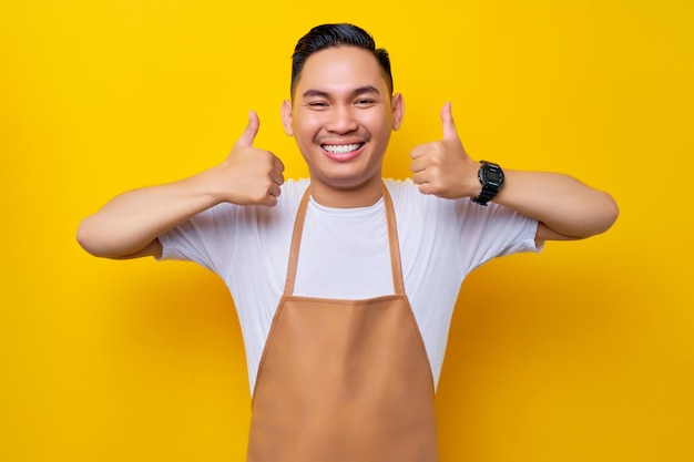 Funcionário de barista jovem asiático sorridente usando avental marrom trabalhando em cafeteria Olhando para a câmera mostrando polegares para cima em aprovação e recomendando em fundo amarelo Conceito de inicialização de pequenas empresas