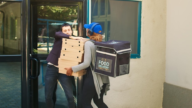 Funcionário de fastfood segurando uma pilha de caixas de pizza para entregar o pedido de almoço ao cliente, serviço de entrega de comida para viagem. Cliente em ligação telefônica recebendo pacotes de refeição do correio da pizzaria na porta da frente.