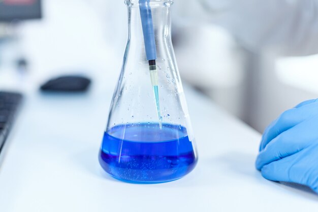 Funcionário de laboratório científico examinando o líquido em frasco de laboratório usando conta-gotas. Químico profissional sênior usando pippete com solução azul para testes de microbiologia.