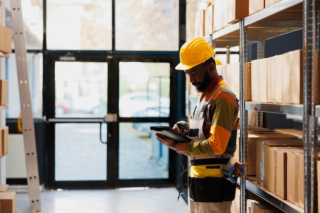 Funcionário do armazém afro-americano verificando o produto digitalizado no tablet digital. Homem gerente de armazém industrial segurando o scanner de código de barras e usando o aplicativo de software de inventário de estoque