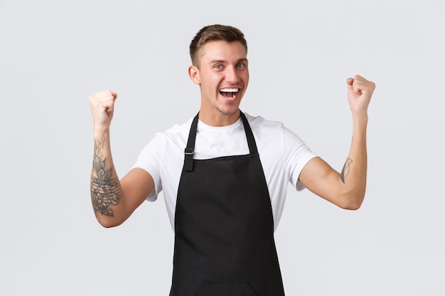 Funcionários, mercearias e conceito de cafeteria. feliz e satisfeito, sorrindo bonito barista, garçom de avental preto, punho erguido de alegria, vitória triunfante ou ótimas notícias positivas