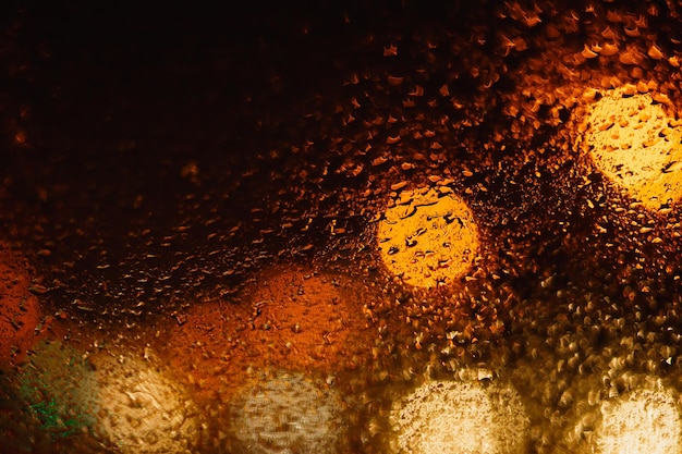 Fundo abstrato escuro com bokeh laranja e amarelo brilhante com gotas de chuva na rua à noite através de vidro, janela