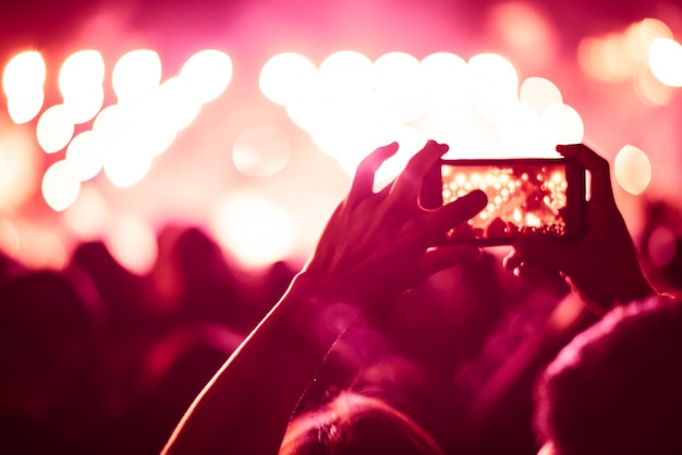 Foto fundo colorido embaçado da mão, segurando o telefone inteligente para tirar fotos pessoas lotadas em concerto.