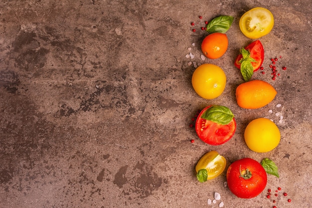 Fundo culinário com tomates maduros e folhas de manjericão. Vários vegetais coloridos, sal marinho e pimenta rosa em um fundo de pedra escura de concreto, vista superior