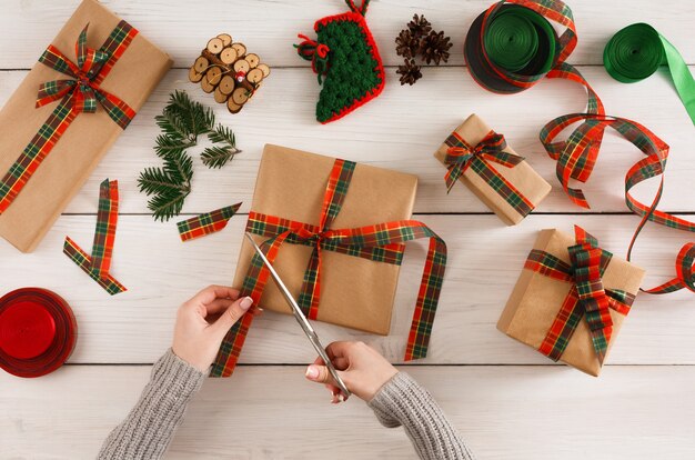 Fundo de embrulho de presente. Vista superior nas mãos femininas embrulhe o presente de Natal em papel artesanal, decore com fita de cetim na mesa de madeira rústica branca bagunçada. Conceito de férias de inverno.