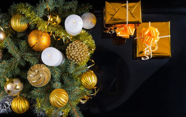 Fundo de Natal com tema dourado com dois presentes decorativos, velas e um arranjo de enfeites e galhos de pinheiro, vista aérea em preto com copyspace