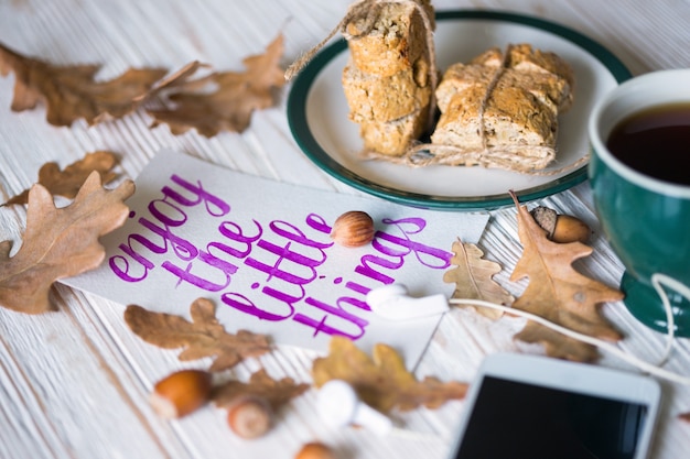 Fundo de outono bonito - xícara de chá, biscotti, folhas de outono e folha com uma citação