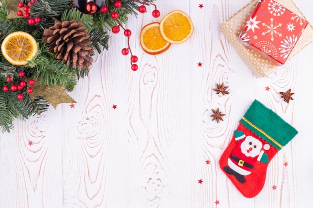Fundo festivo de natal com uma meia de feltro vermelho com a imagem de cones de ramos de abeto de papai noel e presentes de brinquedos de natal laranjas secas em uma mesa de madeira rústica branca Espaço de cópia de vista superior