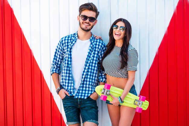 Funky paar. Fröhliche junge Frau, die Skateboard hält und sich mit ihrem Freund verbindet, während sie im Freien steht